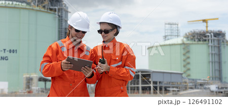 Professional engineer wearing orange suit and white hard hat stands using tablet in at liquefied natural gas facility industrial structures petroleum. Survey planning for gas transmission projects 126491102