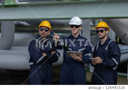 Three construction workers discuss plans while overseeing pipelines at an industrial facility. They are dressed in safety gear and focused on their work in bright daylight. Three construction workers discuss plans while overseeing pipelines at an industrial facility. They are dressed in safety gear and focused on their work in bright daylight. 126491328