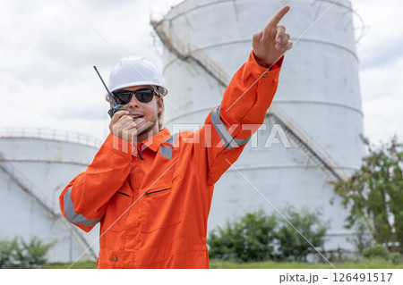 Man professional engineer technician in orange jumpsuit and white hard hat through Walkie Talkie radio while gesturing Pointing forward. Working at tank refining oil petrochemical industry factory. Man professional engineer technician in orange jumpsuit and white hard hat through Walkie Talkie radio while gesturing Pointing forward. Working at tank refining oil petrochemical industry factory. 126491517