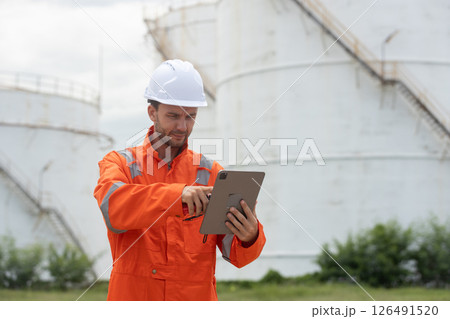 Worker in safety gear using tablet near industrial storage oil tanks refining petrochemical industry factory. 126491520