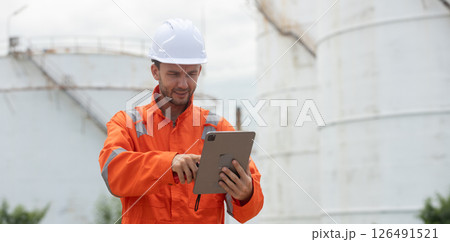 Worker in safety gear using tablet near industrial storage oil tanks refining petrochemical industry factory. 126491521