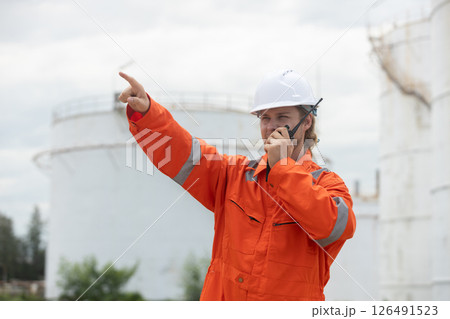 worker in orange safety suit communicates via radio, directing activities at an industrial site with storage tanks. 126491523