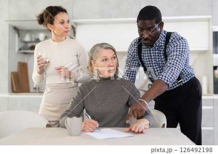 Old woman compiling testament while middle-aged man and woman competing with each other in the kitchen 126492198