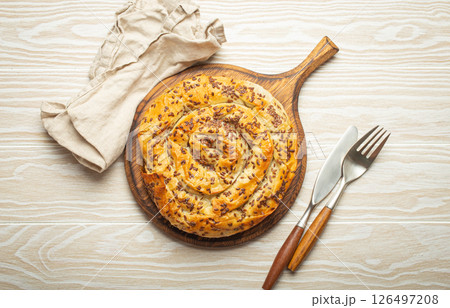 Burek made of filo dough with filling on cutting board, white wooden rustic background top view. Traditional savoury spiral pie of Balkans, Middle East and Central Asia Burek made of filo dough with filling on cutting board, white wooden rustic background top view. Traditional savoury spiral pie of Balkans, Middle East and Central Asia 126497208