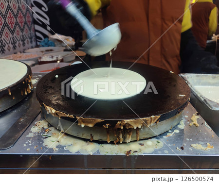 Person pouring ladle of batter into round pan for frying pancakes. Cook making pancakes from fried dough. Frying pancakes outdoors. Cooking street food at festival. Cooking meals on street Person pouring ladle of batter into round pan for frying pancakes. Cook making pancakes from fried dough. Frying pancakes outdoors. Cooking street food at festival. Cooking meals on street 126500574