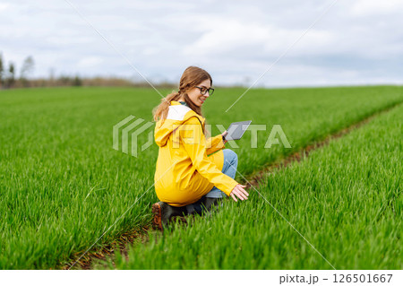 Young farmer woman in a green field, intently using a tablet. She wears a yellow jacket and glasses Young farmer woman in a green field, intently using a tablet. She wears a yellow jacket and glasses 126501667