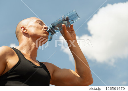 Man in athletic wear is seen drinking water from bottle outdoors, against cloudy blue sky background. Summer heat weather. Thirst. Close up. Man in athletic wear is seen drinking water from bottle outdoors, against cloudy blue sky background. Summer heat weather. Thirst. Close up. 126501670