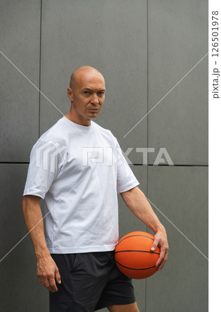 Portrait of handsome sporty basketball player in white t-shirt holding ball and looking at him near the gray wall. Vertical format. 126501978