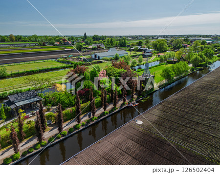 Aerial view of a lush countryside garden surrounded by canals and farmlands, with a cozy house and colorful trees, set in a peaceful rural landscape under a clear blue sky. 126502404