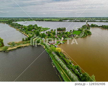Aerial view of a scenic lakeside village with narrow peninsulas, green fields, and houses surrounded by calm waters under a cloudy sky in the Dutch countryside. Aerial view of a scenic lakeside village with narrow peninsulas, green fields, and houses surrounded by calm waters under a cloudy sky in the Dutch countryside. 126502406