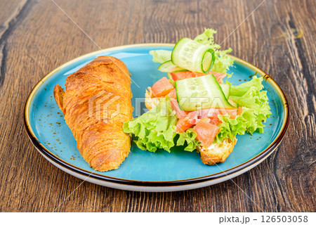 On half of a croissant are ingredients for filling of lettuce leaves, pieces of red fish and cucumbers. On half of a croissant are ingredients for filling of lettuce leaves, pieces of red fish and cucumbers. 126503058