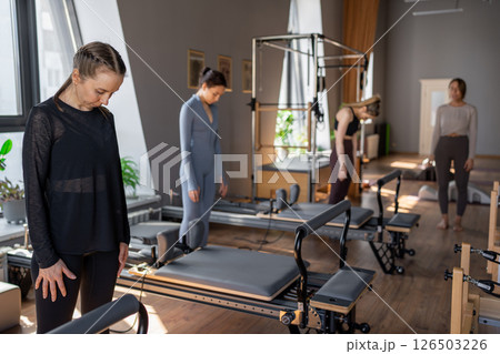 Young caucasian woman doing roll down exercise in a pilates bright studio. Their workout focuses on core strength and flexibility in a modern fitness environment. 126503226