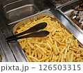 Crispy golden french fries resting in metal buffet tray, ready for serving with black tongs amid food station lineup 126503315