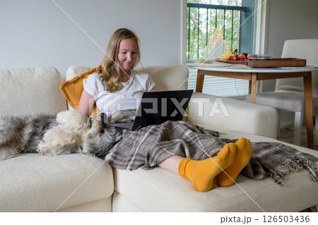 Woman with bright socks sitting on cream sofa with blanket and dog and using laptop Woman with bright socks sitting on cream sofa with blanket and dog and using laptop 126503436