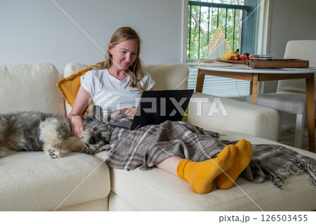 Woman with bright socks sitting on cream sofa with blanket and dog and using laptop Woman with bright socks sitting on cream sofa with blanket and dog and using laptop 126503455