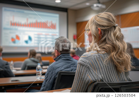 Group of employees attend a training session in a conference room. Charts and graphs are displayed on a screen in the background, while the attendees focus intently on the presentation 126503504