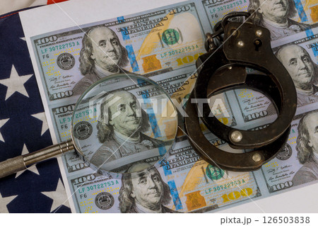 Handcuffs rest top multiple US hundred dollar bills while magnifying glass focuses on one bill serious mood evokes an investigative atmosphere. 126503838