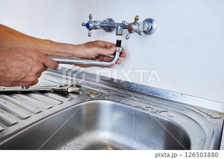 Person is installing faucet spout into wall-mounted tap above kitchen sink while using hands and water is visible on stainless steel surface. 126503880