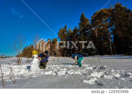 Husband and wife build snowman, adding final touches, placing bucket on its head, enjoying sunny winter day, wearing warm clothes and gloves. 126503896