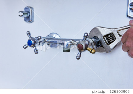 Removing old faucet for replacement with new one, worn metal tap with blue and red handles is being loosened with adjustable wrench on white wall. 126503903
