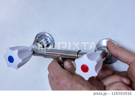 Hands are installing new two handle faucet onto wall outlets, plumber tightens metal fixture with white handles to exposed threaded connectors. 126503916