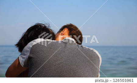 Happy couple hugs during sitting on wooden log on sandy beach. Romantic man kissing his beloved near coastline. Carefree lovers spending time together at resort. Concept of summer vacation or holiday 126503961