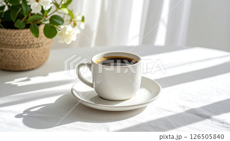 A white coffee cup and saucer on a sunlit table with a plant and curtains in the background. 126505840
