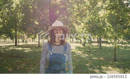 Young woman stands joyfully in a park filled with trees and fallen leaves, wearing a hat and overalls on a warm, sunny afternoon in autumn. Young woman stands joyfully in a park filled with trees and fallen leaves, wearing a hat and overalls on a warm, sunny afternoon in autumn. 126505913