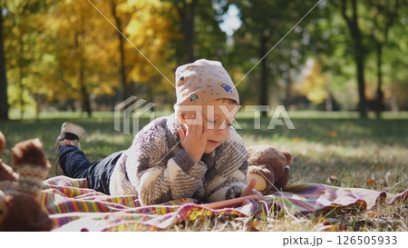 A young child relaxes on a blanket in a park, resting their chin on their hand, surrounded by plush toys and vibrant autumn leaves under bright sunlight. 126505933