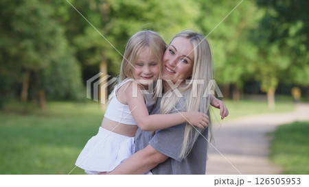 A smiling mother holds her young daughter in a sunny park. They enjoy a playful moment, surrounded by trees and fresh grass on a warm afternoon. 126505953