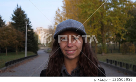 A young woman with curly hair wears a gray cap, enjoying a peaceful walk in a leafy park as the sun begins to set, creating a serene atmosphere. 126505959