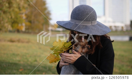 A woman embraces her small dog while holding colorful autumn leaves. Bright sunlight filters through the trees in a vibrant city park during the fall season. 126505978