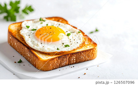 Fried egg on toasted bread with pepper and herbs, served on white paper on a bright surface. Fried egg on toasted bread with pepper and herbs, served on white paper on a bright surface. 126507096