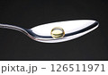 A single yellow softgel capsule sits on a silver spoon against a dark background, focusing on health and medicine. 126511971