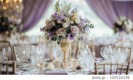 Elegant indoor wedding dining table with floral arrangements in shades of purple, lavender and pink, set in a grand room with mirrors and chandeliers. 126516198