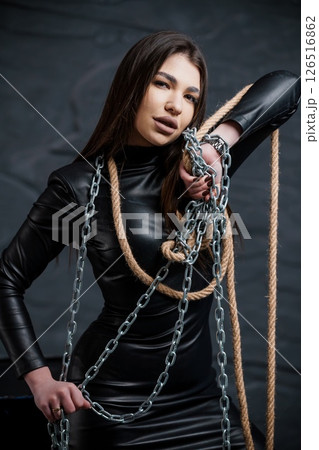 A captivating young woman with striking features is bound with chains and rope in a dark, dramatic studio setting, wearing a sleek black leather dress 126516862