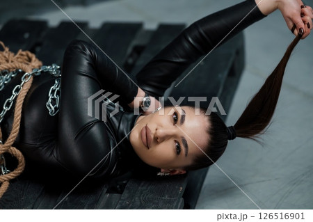 A striking woman with a sleek ponytail and leather top lies confidently on a wooden pallet with chains, exuding a strong and rebellious aesthetic in a dimly lit industrial setting 126516901