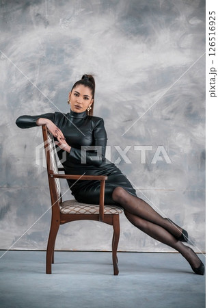A striking woman with dark hair in a sleek black leather dress and sheer black tights sits poised on a vintage wooden chair against a textured grey backdrop 126516925
