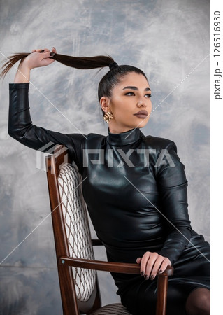 A captivating portrait of a young woman with a sleek ponytail, striking a pose with confidence in a black leather dress, seated on a wooden chair 126516930