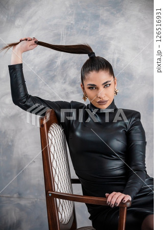 Striking woman with long dark hair in a high ponytail poses confidently in a black leather dress, seated on a vintage chair against a textured grey backdrop Striking woman with long dark hair in a high ponytail poses confidently in a black leather dress, seated on a vintage chair against a textured grey backdrop 126516931