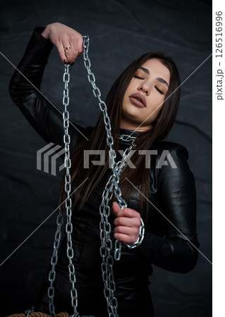 A young woman with long dark hair gracefully holds a prominent silver chain with closed eyes against a dark, textured backdrop, embodying themes of captivity and strength 126516996