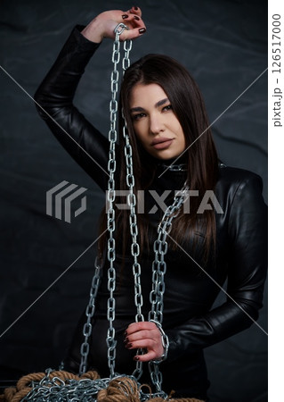 A striking young woman with long dark hair poses intensely while holding and draped in silver chains against a dark, textured backdrop, embodying a sense of power 126517000