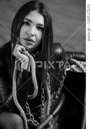 A striking black and white portrait of a young woman with long dark hair sitting in a leather chair with a rope and chain detail for a dramatic look 126517023
