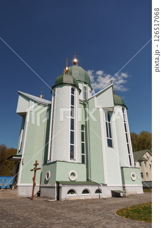 Exterior view of a modern light green Orthodox church with golden domes under a bright blue sky featuring a cross and surrounding trees 126517068