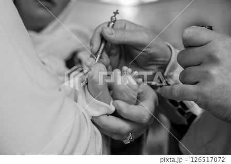 A close-up black and white photograph depicts a solemn baptism ceremony a priest anoints the foot of a newborn baby with holy oil using a small silver vessel signifying spiritual initiation 126517072