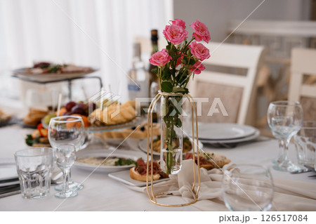 Elegant table setting with a vase of pink roses, appetizers and wine glasses, creating a celebratory atmosphere in a bright, modern dining room 126517084
