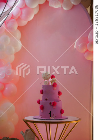 A charming First Birthday celebration featuring a three-tiered pink cake decorated with lanterns and a cute animal figurine against a vibrant balloon backdrop in a pink setting A charming First Birthday celebration featuring a three-tiered pink cake decorated with lanterns and a cute animal figurine against a vibrant balloon backdrop in a pink setting 126517086