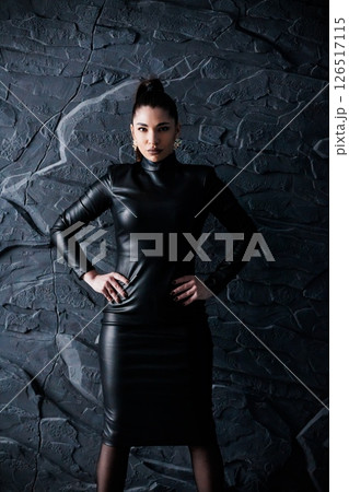 Dramatic portrait of a confident woman in a sleek black leather dress posing against a dark textured stone wall, embodying power and sophistication 126517115