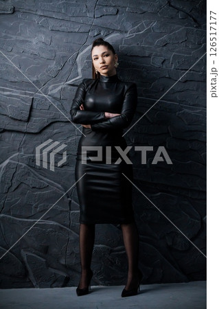 Dramatic portrait of a young Hispanic woman in a sleek black leather dress standing confidently with crossed arms against a textured stone wall backdrop 126517177
