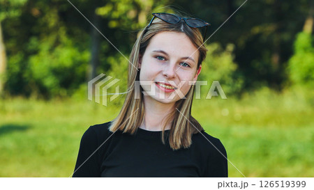 Smiling blonde teenage student in a black t shirt and sunglasses posing in a green park 126519399